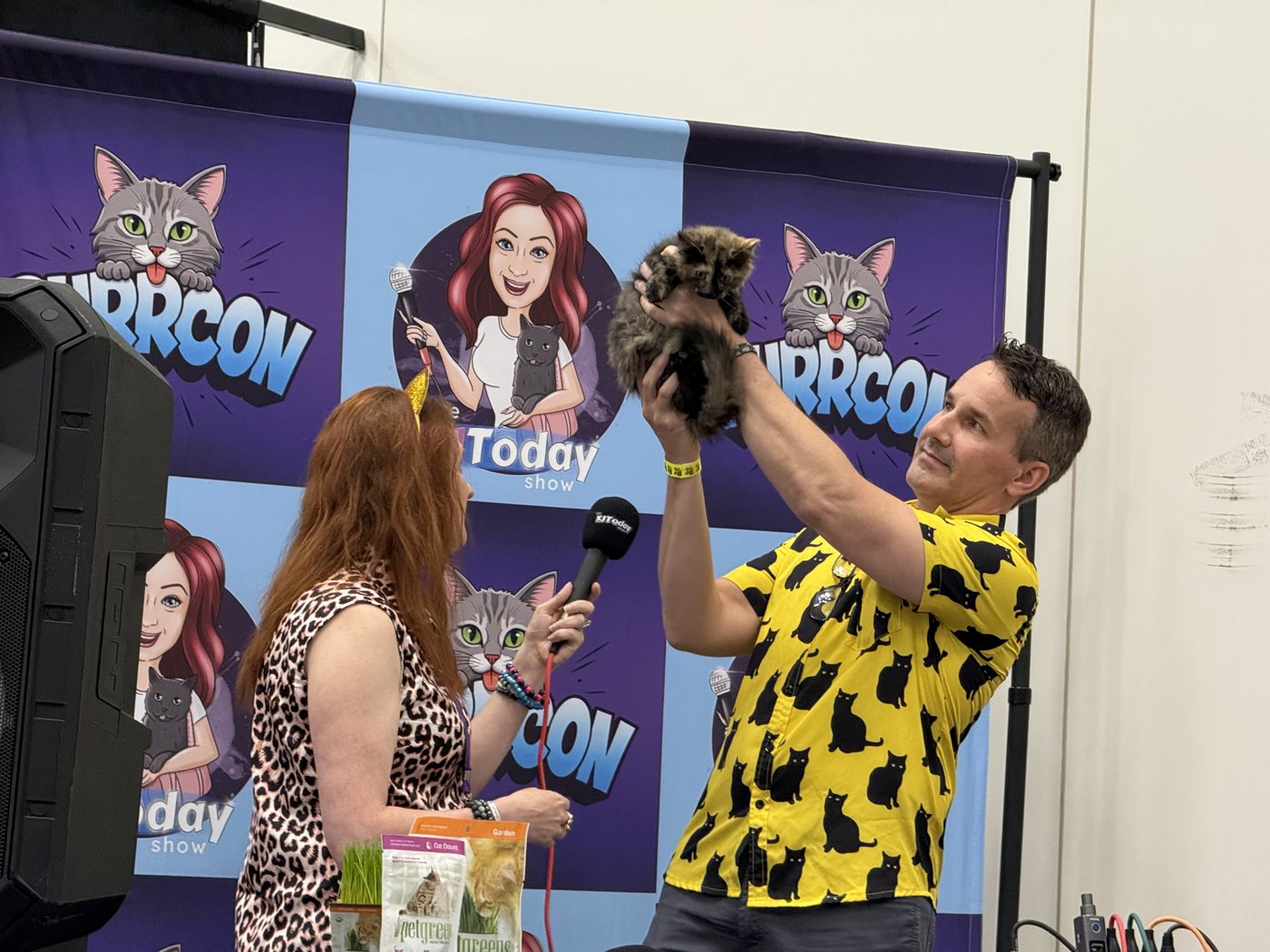 Dr. Matt McGlasson holding up a fluffy kitten on the PurrCon stage while KJ holds the microphone