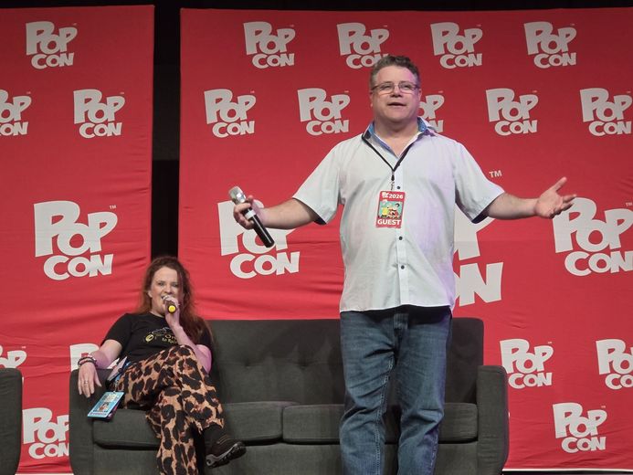 Speaker with a microphone on the PopCon stage beside a seated guest